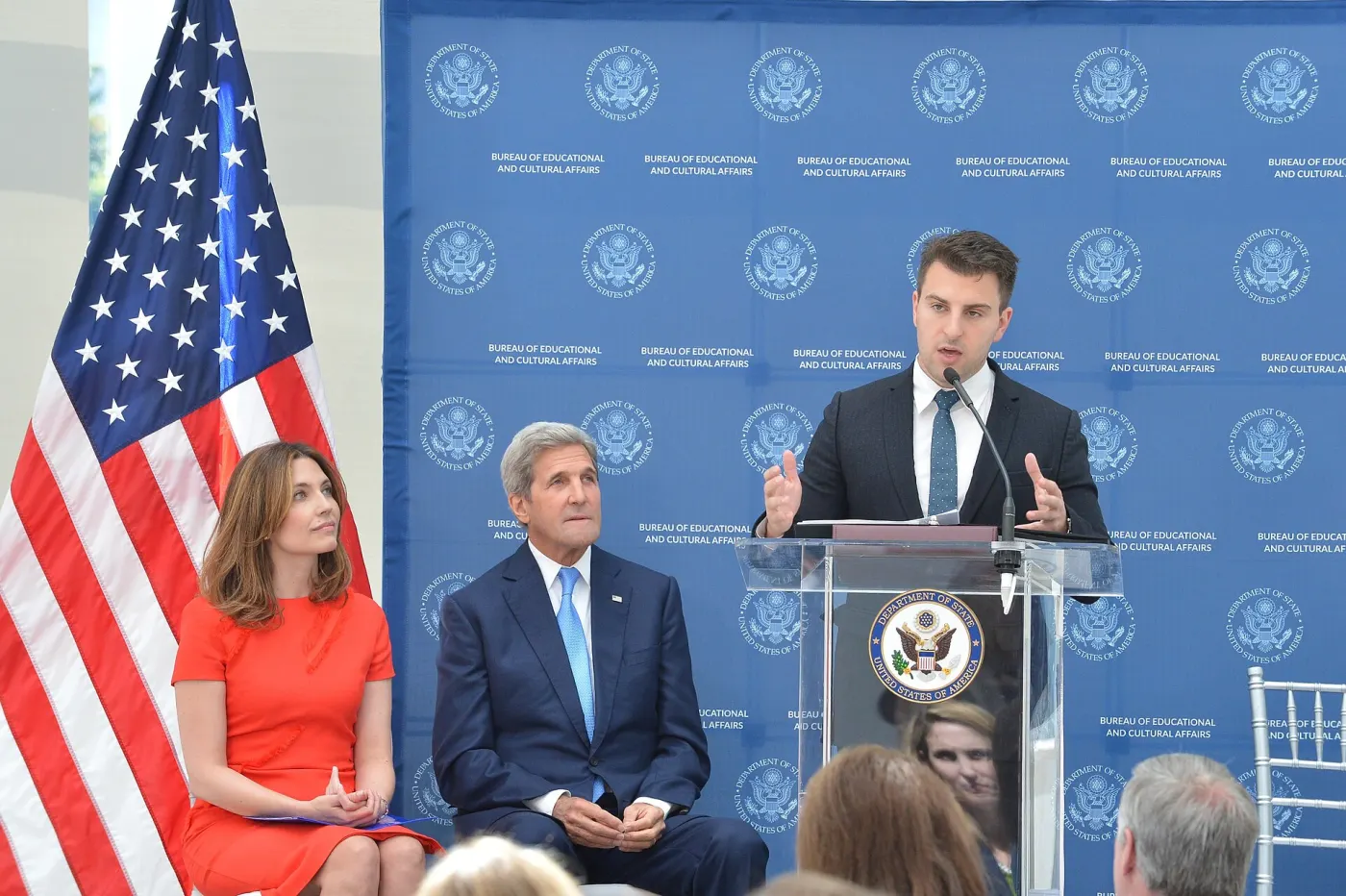 Chesky speaking at State Department podium with Kerry seated