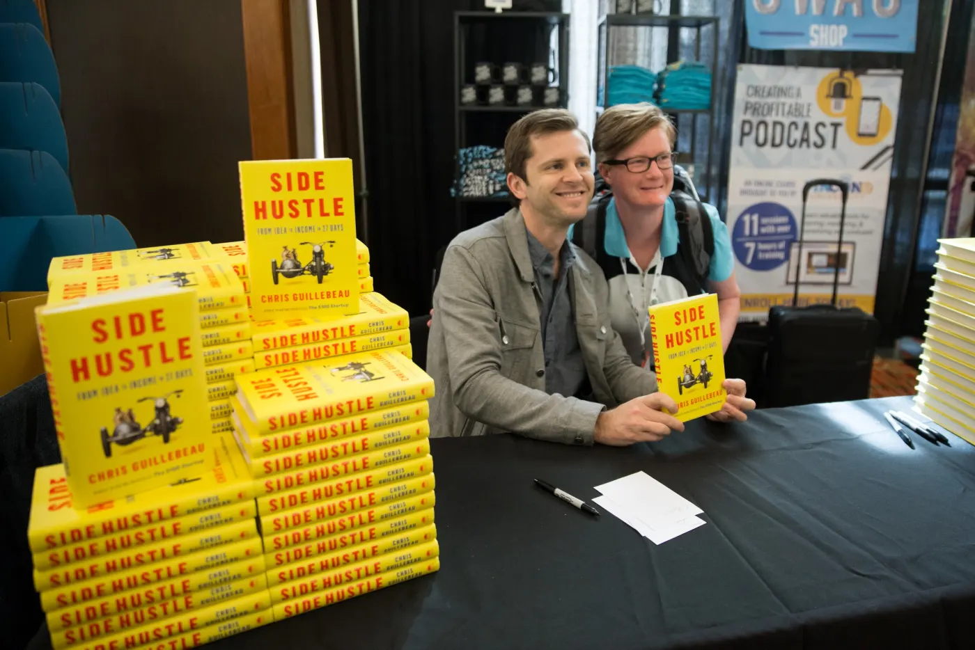 Guillebeau with fan and stacks of Side Hustle at event swag area