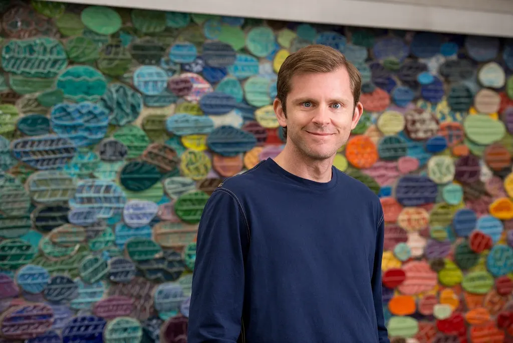 Smiling Guillebeau portrait in a navy sweater in front of a colorful mosaic wall