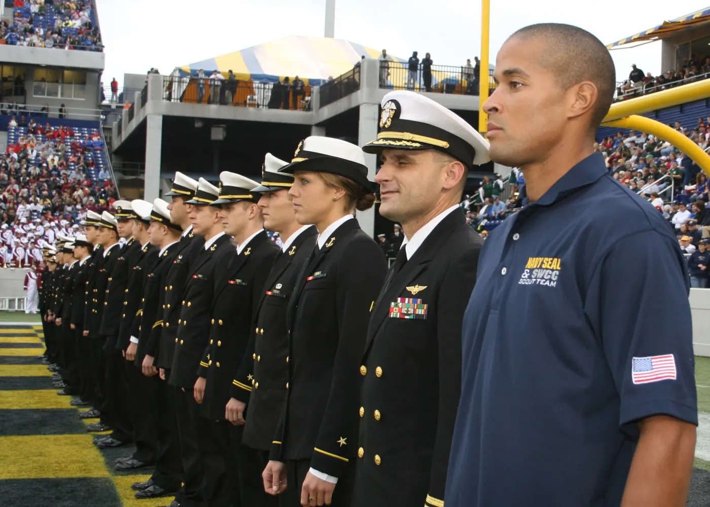 Goggins in a Navy polo shirt standing beside uniformed Navy personnel at a stadium