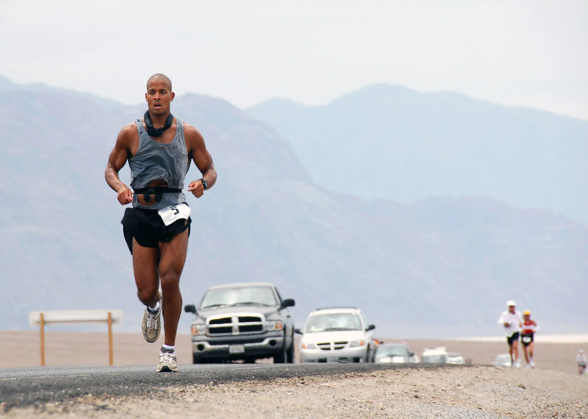 Goggins running on a desert road with mountains behind him and crew vehicles following