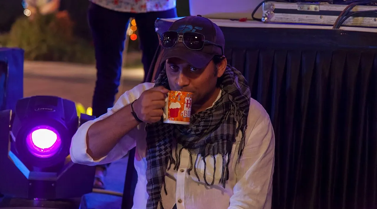Shah at a night event in white shirt and scarf, sipping from a birthday mug