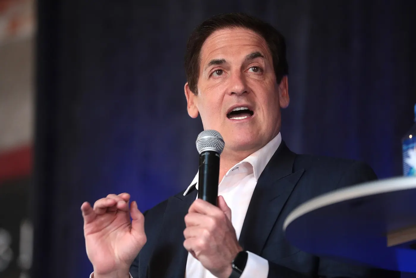 Mark Cuban speaking into a microphone on a stage, open-collar white shirt and dark blazer