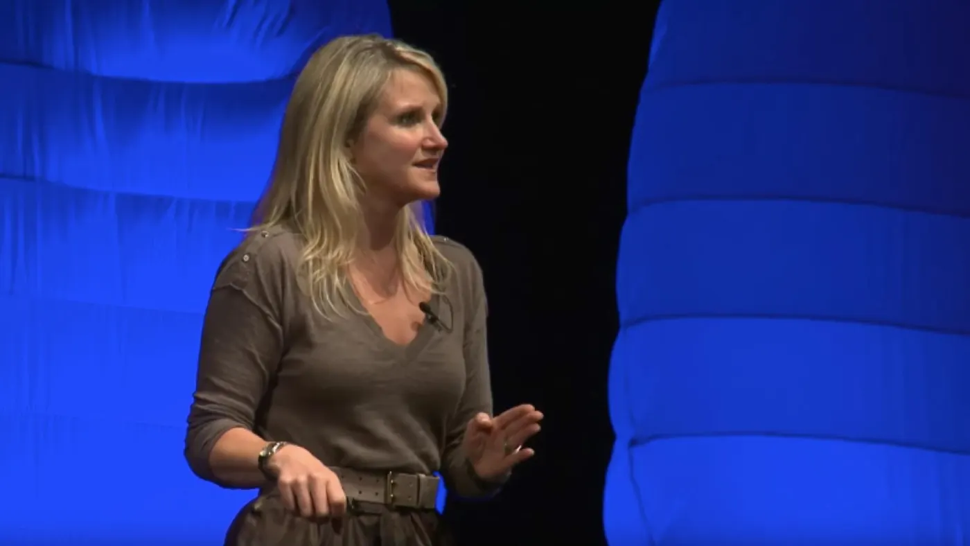Mel Robbins speaking on a dark stage lit in blue, in a gray top