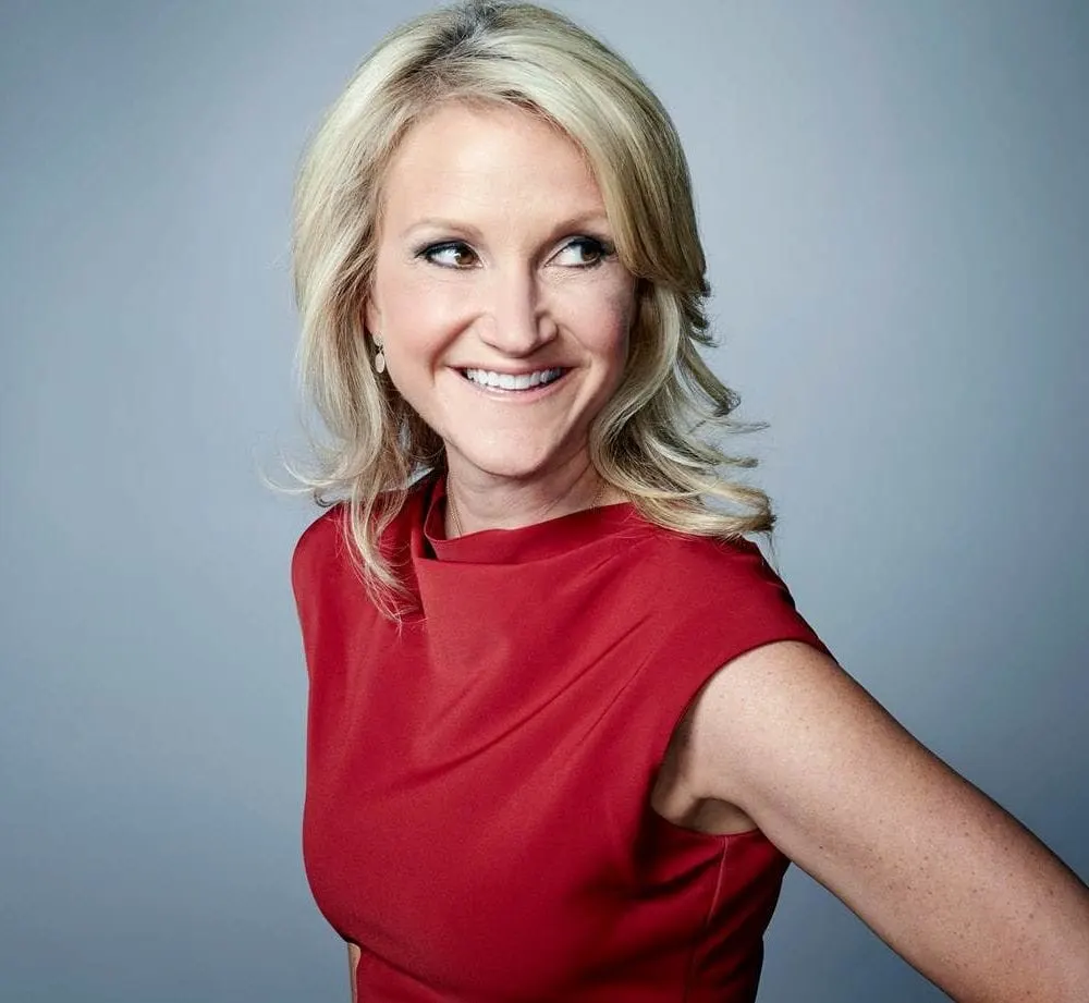 Mel Robbins smiling in a red sleeveless top against a blue-gray studio background