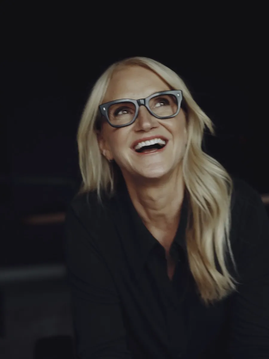 Mel Robbins laughing with her head tilted back, wearing dark-rimmed glasses and a black top