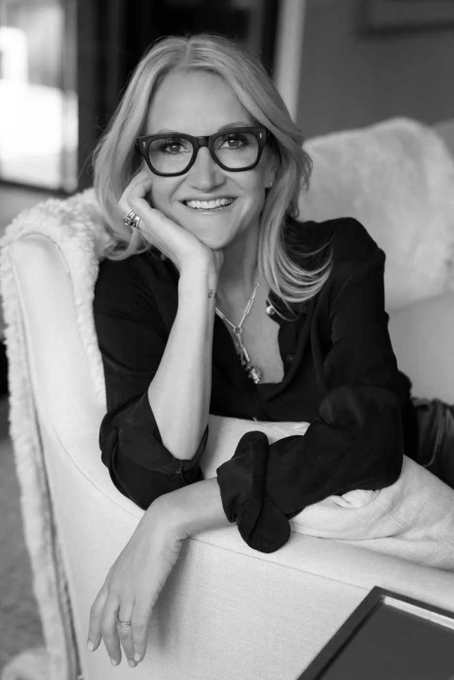Mel Robbins in black and white, glasses on, chin resting on her hand in a white armchair