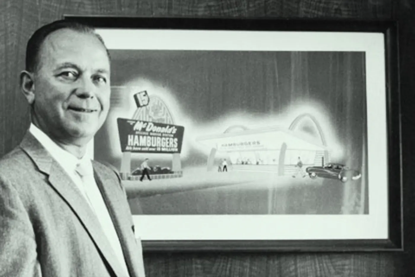Ray Kroc in a light suit beside a framed picture of an early McDonald's