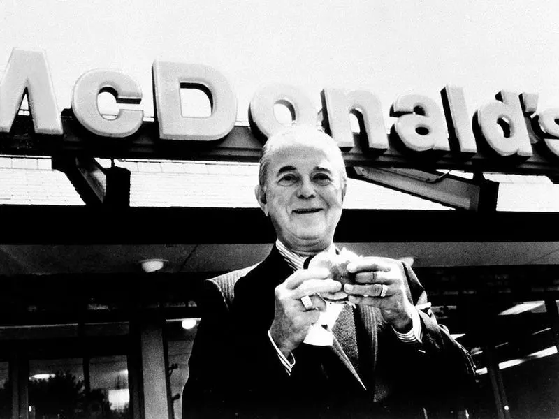 Ray Kroc smiling and holding a burger beneath a large McDonald's sign