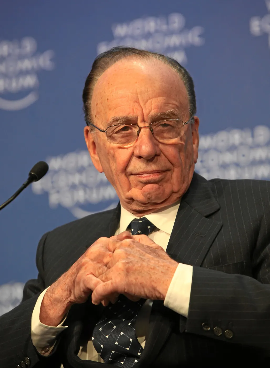 World Economic Forum seat, hands clasped, pinstripe suit