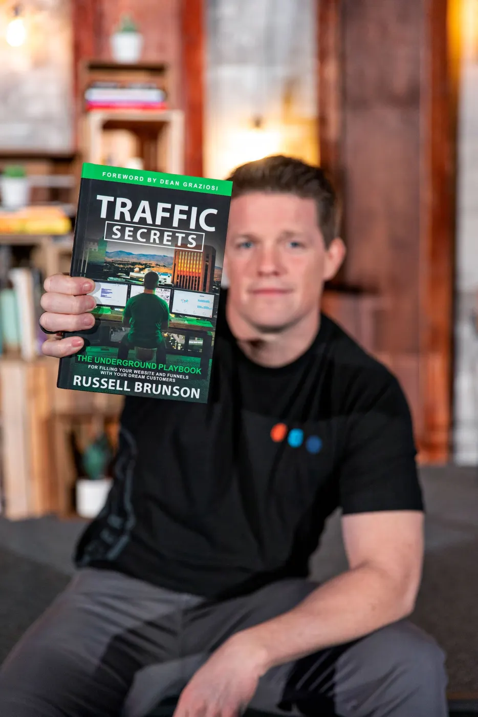 Russell Brunson in a black T-shirt holding up a copy of Traffic Secrets in front of bookshelves