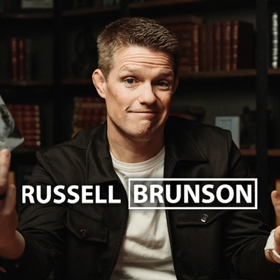 Russell Brunson in a dark jacket and white T-shirt, grinning in front of bookshelves, with his name in large letters