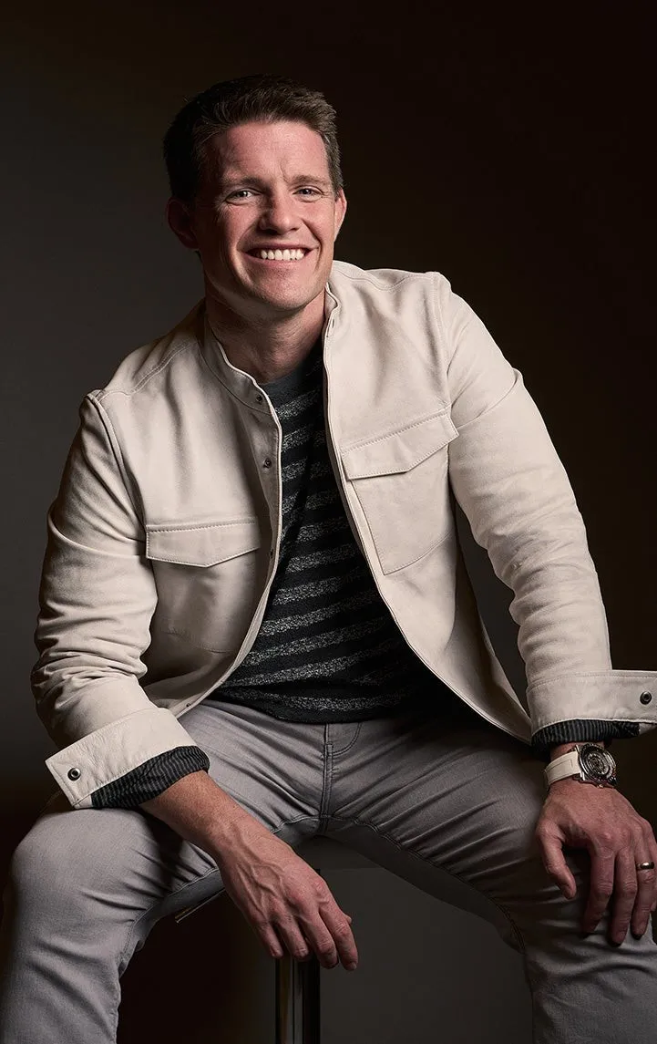 Russell Brunson seated on a stool in a cream jacket and striped shirt against a dark background