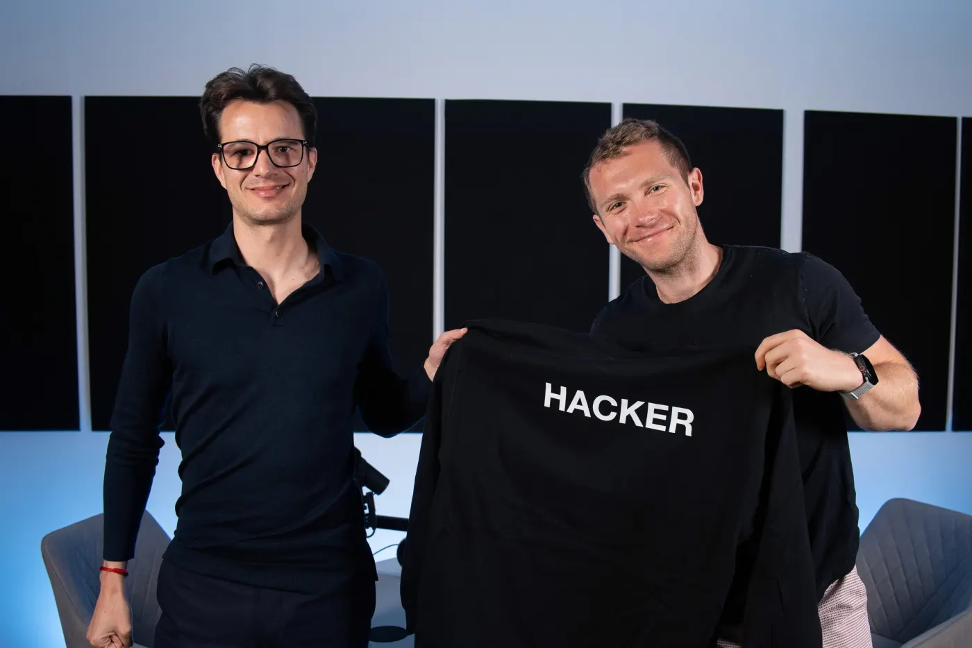 Hacker smiling next to podcast host Harry Stebbings, who holds up a black shirt that says "HACKER"