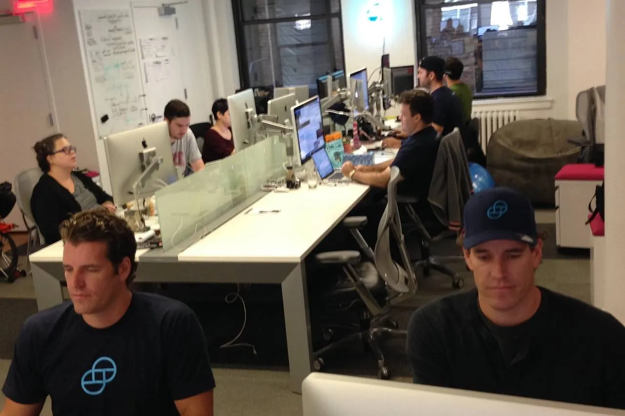 A Winklevoss twin at a desk in a busy office with Gemini logo shirts